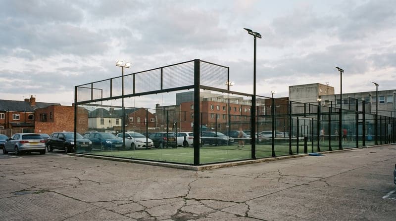 Floodlit outdoor padel courts with glass end walls and mesh fencing beside a car park