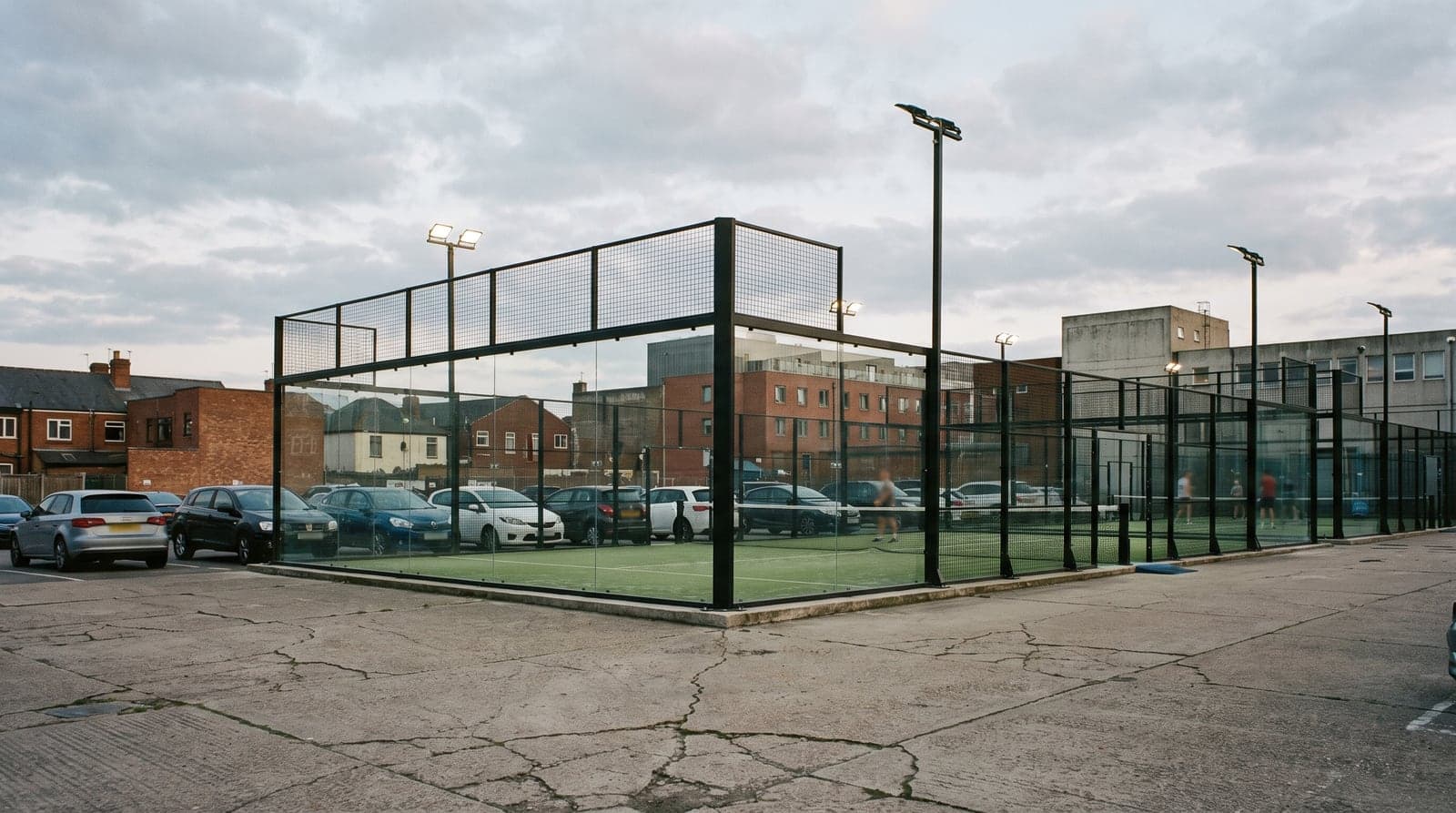 Floodlit outdoor padel courts with glass end walls and mesh fencing beside a car park