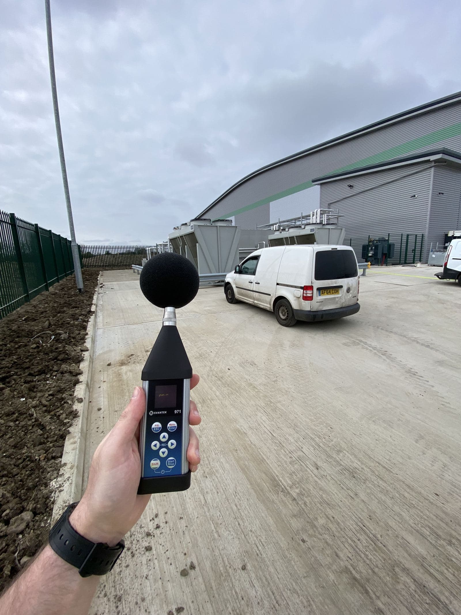 An engineer holding a Class 1 sound level meter beside an industrial unit with rooftop plant